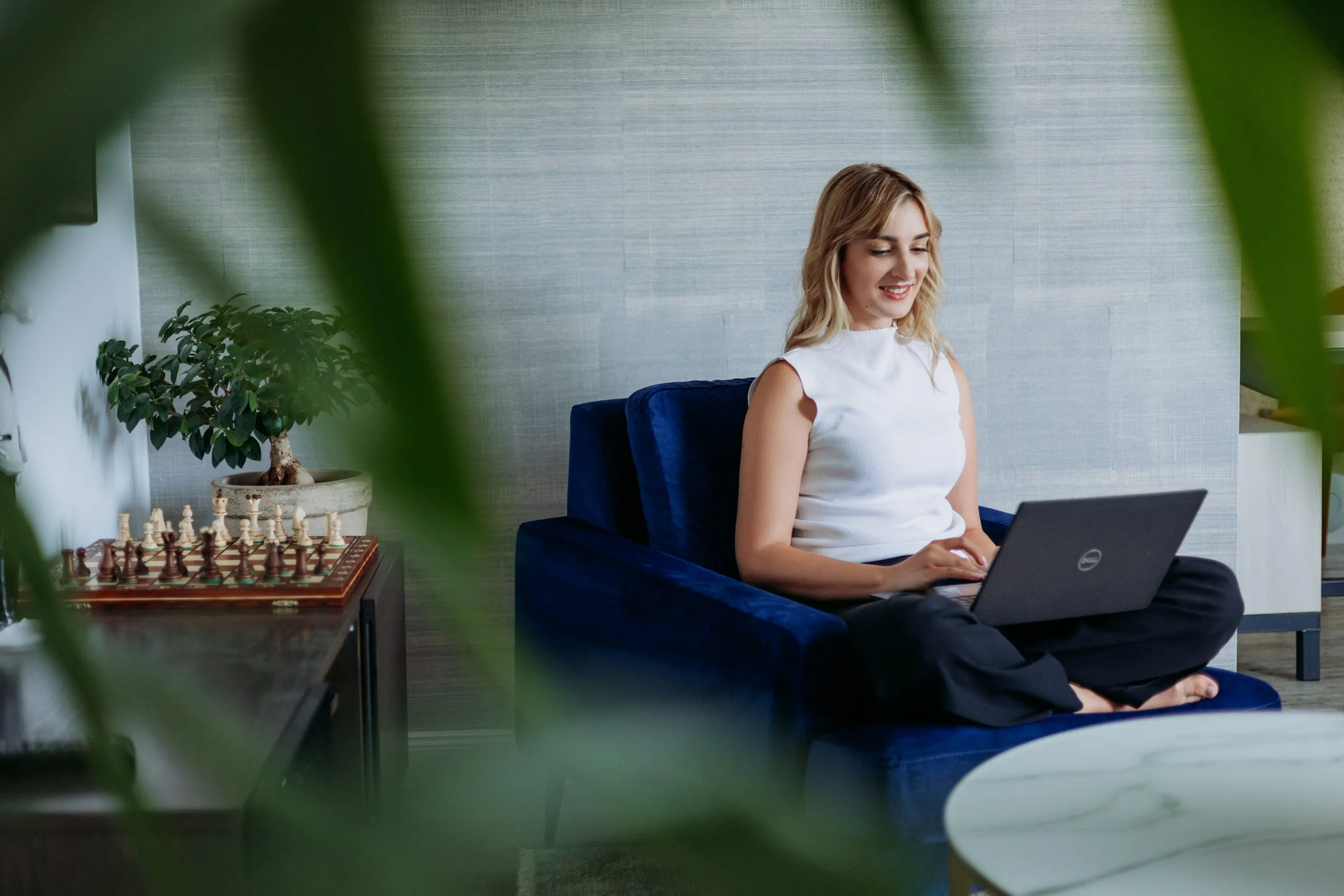 Miruna Zamfiriu working from a blue armchair beside a chess board and a bonsai